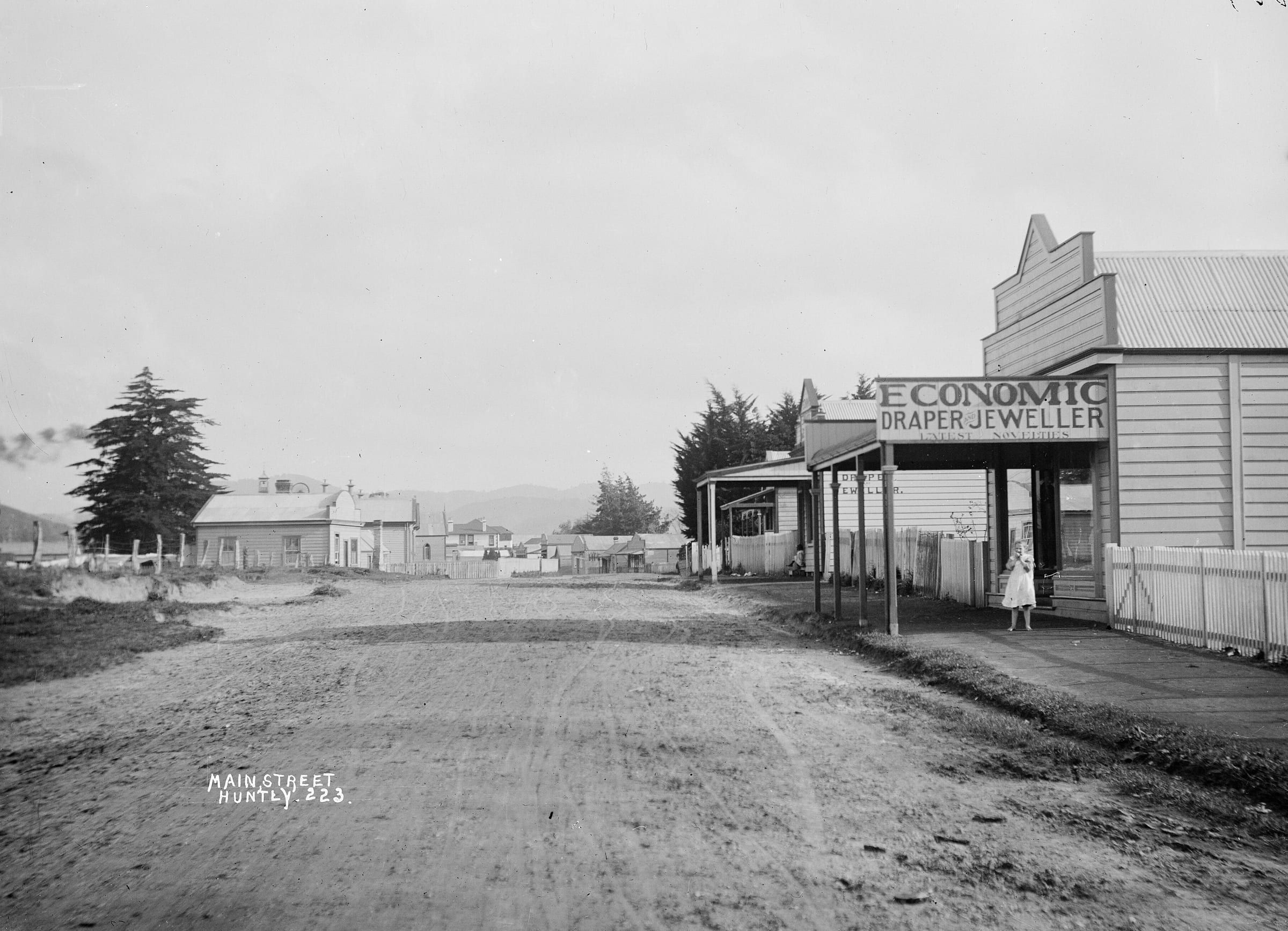 Black and white photo of Main Street, Huntly, in the 1910s: a dirt road, timber shops with verandahs and a sign for an Economic Draper and Jeweller.