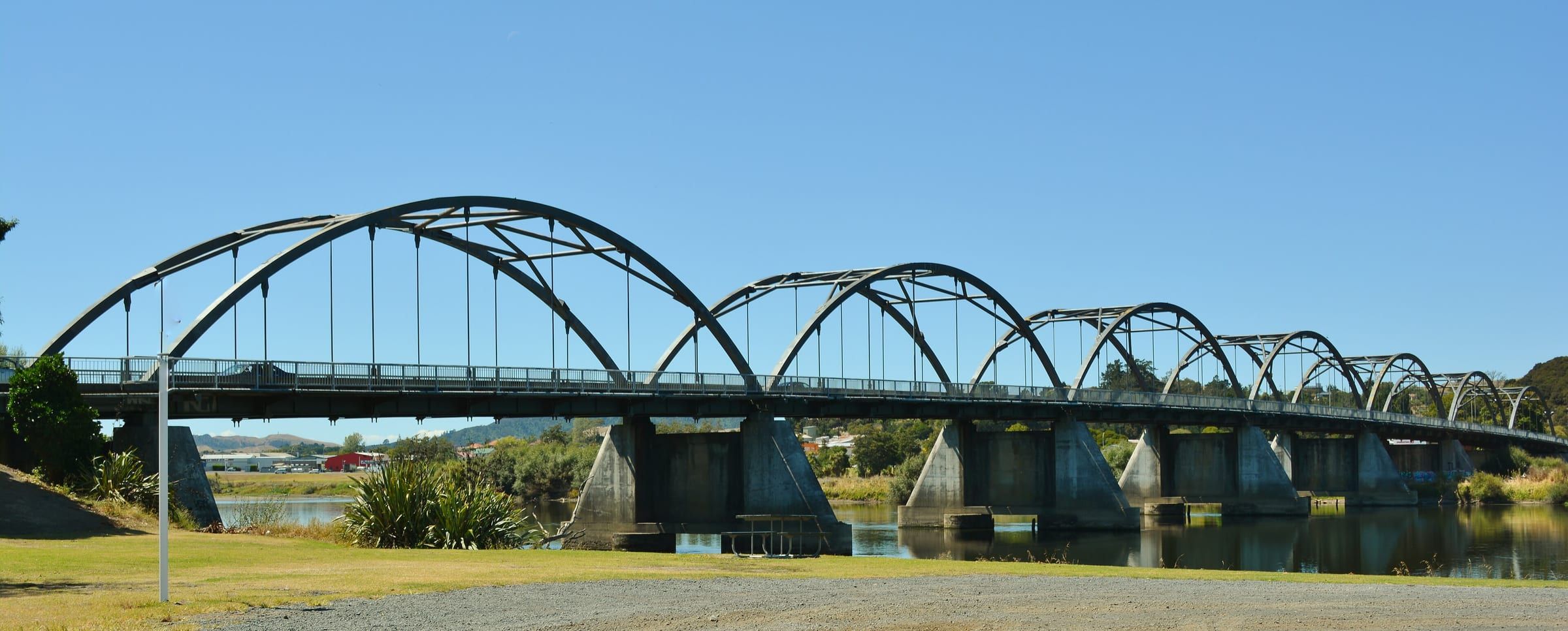 The Tainui Bridge, a row of steel arches, spanning the Waikato River at Huntly under a blue sky.